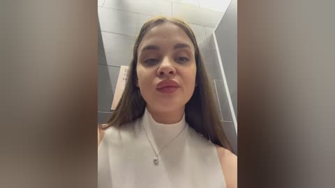 Video of a young woman with straight brown hair, wearing a white sleeveless top, standing in a tiled bathroom with light beige walls and a silver towel rack.