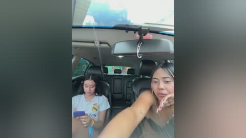 Video of a young woman with dark hair, wearing a sleeveless top, leaning on the car window. She holds a smartphone. In the background, a girl with long hair in a white t-shirt looks at her phone.