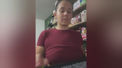 Video of a short, light-skinned woman with dark hair, wearing a maroon T-shirt, standing in a supermarket aisle. Shelves are filled with colorful snacks and beverages, and the background is blurred.