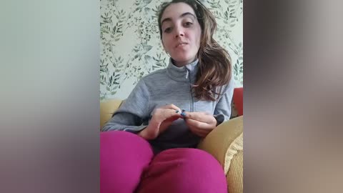 A video of a young woman with light skin, brown hair, and a light gray zip-up sweater, sitting on a mustard-yellow couch with magenta leggings, surrounded by a patterned wallpaper.