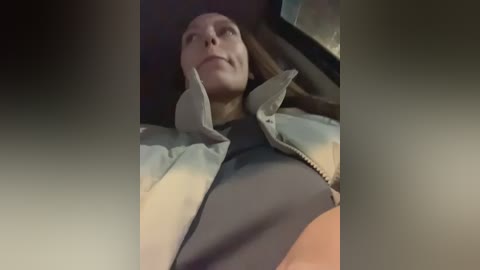 A video of a woman in a car, wearing a gray jacket over a black shirt, lying on her back with her head slightly elevated. The background is blurry, indicating motion.