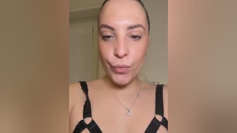 A close-up video of a light-skinned woman with a straight, dark-haired ponytail, wearing black strappy lingerie and a silver necklace. Her face is pouty with slightly raised eyebrows. Background is blurred, indoor setting.