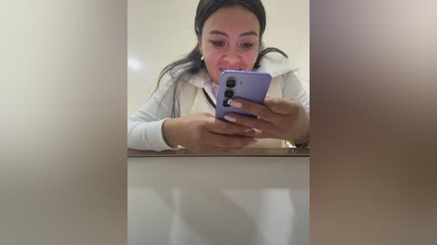 Video of a young woman with medium skin tone and dark hair, wearing a white sweater, intently looking at her purple smartphone in a mirror reflection.
