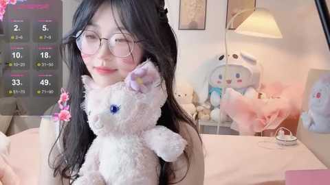 A video of an East Asian woman with long black hair, fair skin, and large glasses, holding a plush white bunny, surrounded by pastel decor, including a white lamp and plush toys.