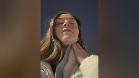 A video of a young woman with long brown hair, closed eyes, and a serene expression, wearing a white sweater. A person's hand is gently touching her neck. The background is a gradient of dark blue to black.