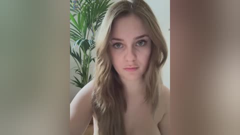 Video of a young Caucasian woman with long, wavy blonde hair, fair skin, and blue eyes, partially nude, in a modern indoor setting with greenery and soft lighting.