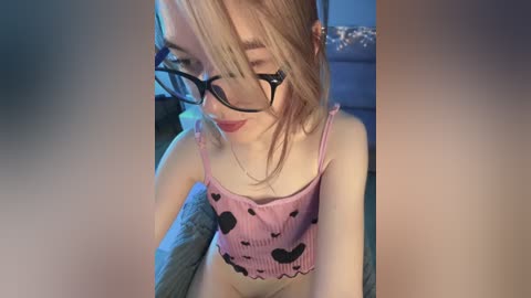 A young woman with fair skin, blonde hair, and glasses sits on a bed, wearing a pink heart-patterned crop top, smiling with closed eyes.