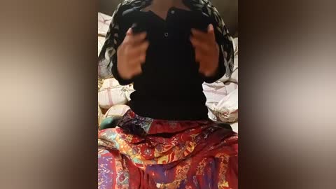 A video captures a woman in a black top, holding her hands in front of her face, seated on a bed with a colorful, patterned blanket and pillows in the background.