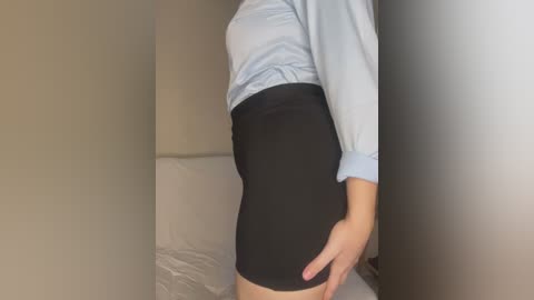 Video of a light-skinned woman in a light blue blouse, black tight-fitting skirt, and white underwear. She stands in a dimly lit room with beige walls and a white bed.