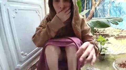 A candid video of a young girl in a brown hoodie, with her hand over her mouth, sitting on a purple skirt. She is in a fenced outdoor area with a green plant and a white door in the background.