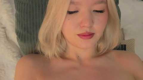 A video of a young Asian woman with light skin and shoulder-length blonde hair, wearing minimal makeup and looking down, against a background of soft, muted pillows.