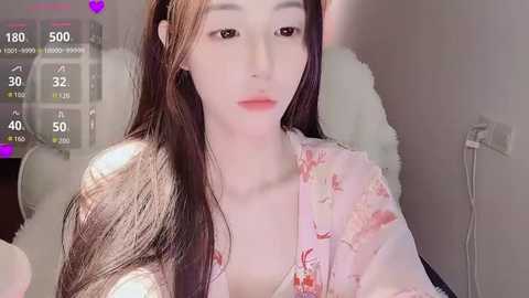 Video of a young Asian woman with long, straight brown hair, light skin, and a slender physique, wearing a floral robe. She has a neutral expression, and the background shows a bedroom with a bed and a white wall.