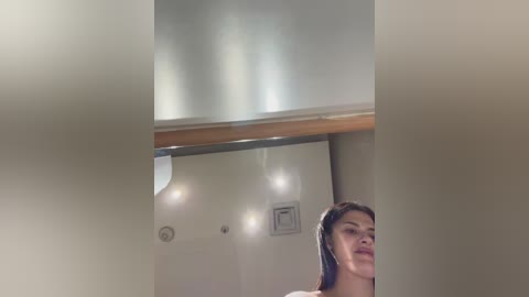 A video of a woman with long, dark hair, wearing a black top, partially visible through a fogged-up window. The background shows a beige wall with a light fixture and a wooden railing.