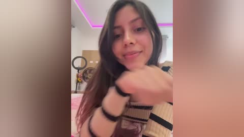 Video of a young woman with long brown hair, wearing a striped sweater, making a \"thumbs-up\" gesture, in a cozy bedroom with pink LED lights.