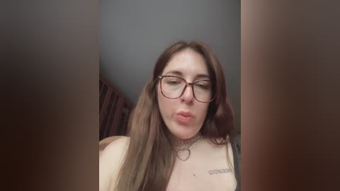 A video of a fair-skinned woman with long brown hair, wearing glasses and a black top, sitting indoors.