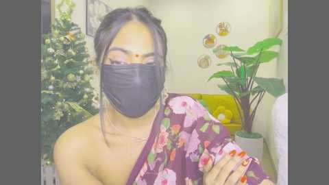 Video of a woman with medium skin tone, wearing a floral kimono, black face mask, and red nail polish, sitting in a room with a Christmas tree, potted plants, and wall art.