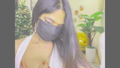 Video of a woman with long, straight black hair, wearing a black face mask, white halter top, and gold earrings, in a room with a Christmas tree, green plant, and yellow couch.