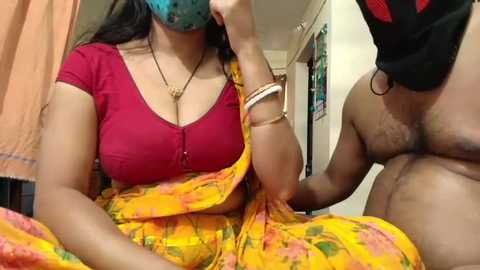 Video of a South Asian woman in a red blouse, yellow sari, and blue mask, sitting on a bed with a man's erect penis visible.