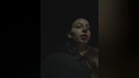 A dimly-lit, grainy video of a woman with fair skin, dark hair, and closed eyes, wearing a black jacket and headphones, against a dark, shadowy background.