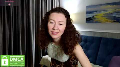Video of a smiling, curly-haired woman in a leopard-print top, sitting on a blue couch, with dark curtains and a painting in the background.