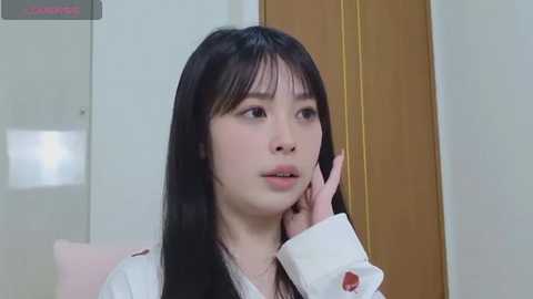 A video of an East Asian woman with long black hair, wearing a white shirt, sitting in a room with a wooden door and light walls. She appears contemplative.