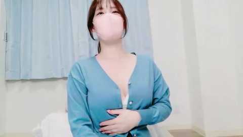 Video of a young Asian woman in a blue cardigan, sitting with a hand on her chest, wearing a surgical mask in a sterile hospital room with light blue curtains.
