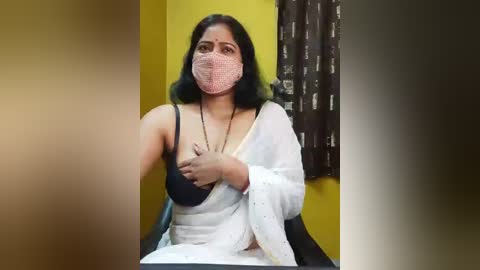 Video of a South Asian woman with medium skin tone, wearing a pink face mask, black bra, white shawl, and black pants, seated indoors against a yellow wall, with a brown curtain in the background.