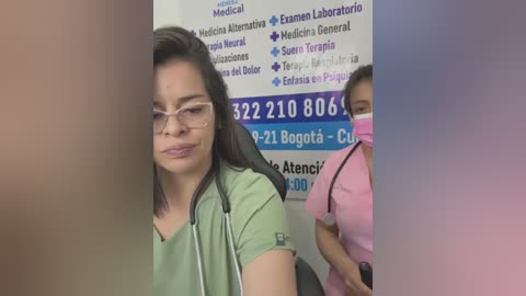Video of a smiling Latina woman with glasses and a stethoscope, wearing a green scrub top. Background includes two medical staff in scrubs, a white wall with a contact information sign.