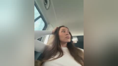 Video of a Caucasian woman with long brown hair, wearing a white top, sitting in a car. She is adjusting her hair with her left hand. The background shows a window and a clock.