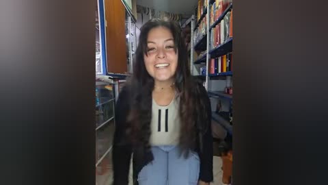 Video of a smiling woman with long, dark hair, wearing a white tank top and light blue jeans, standing in a cluttered bookshelf-lined room.
