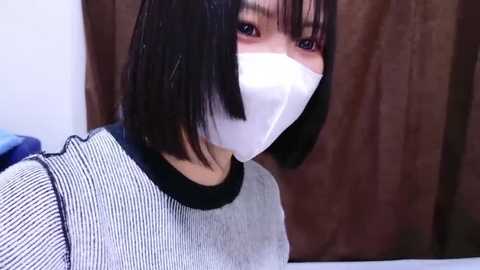 Video of a person with short black hair, wearing a white face mask, gray striped shirt, and dark-rimmed glasses, in a room with brown curtains in the background.