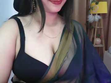 Media: Video of a smiling woman with fair skin and long, dark hair, wearing a black bra, draped in a sheer, multicolored saree. Background shows a lampshade, a bookshelf, and a potted plant.