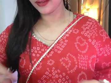 Media: Video of a woman with long black hair, wearing a red saree with white floral patterns and a gold chain, standing indoors near a lamp.