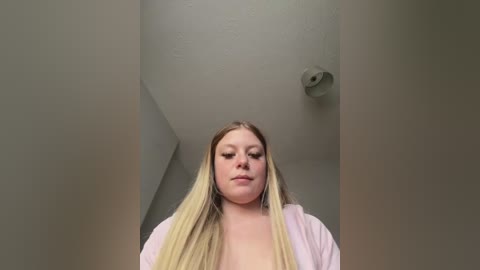 A video of a blonde Caucasian woman with long, straight hair, wearing a light pink robe, standing in a plain, white-walled room with a ceiling light fixture.