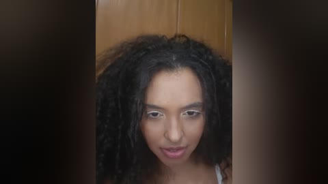 Video of a young woman with a curly black afro, light brown skin, and striking facial features. She has a neutral expression, wearing a white tank top. The background is a wooden door.