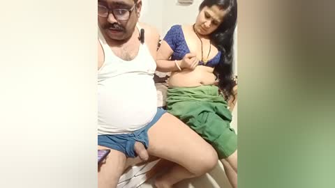 A video captures an overweight man in a white tank top and blue shorts, holding a partially undressed woman in a green saree and blue bra, with long black hair, in a dimly lit room.