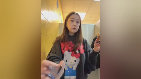 Video of an Asian woman with straight, shoulder-length brown hair wearing a black \"Hello Kitty\" sweatshirt, holding a donut, standing in a yellow-tiled hallway with fluorescent lighting.