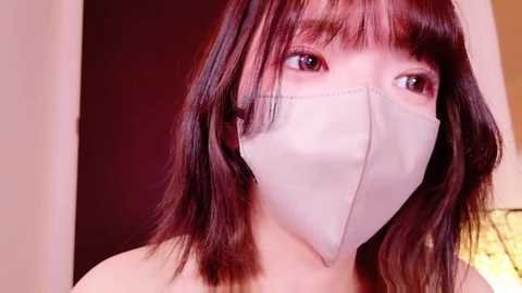Video of an East Asian woman with straight, shoulder-length dark brown hair, wearing a white surgical mask and a beige top. Background is a warm, blurred indoor setting with muted colors.