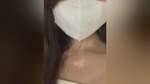 A close-up video of a person wearing a white surgical mask, partially covering their face, with long dark hair visible. The background is blurred, featuring warm tones.