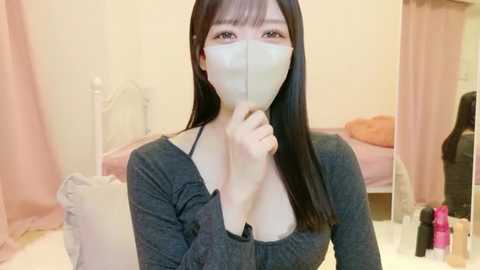 A video of an Asian woman with long black hair, wearing a gray long-sleeve top, a white face mask, and a white handkerchief covering her mouth, sitting on a white bed in a cozy bedroom.