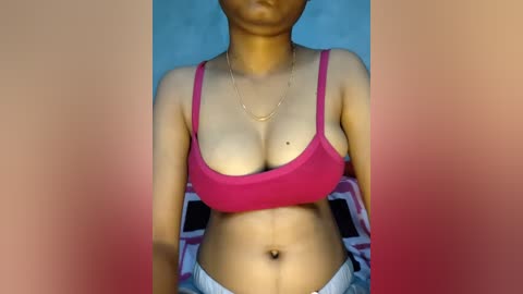Video of a light-skinned woman with a slender physique, wearing a pink sports bra and white shorts. Her face is partially blurred. Background features a bed with a patterned blanket.