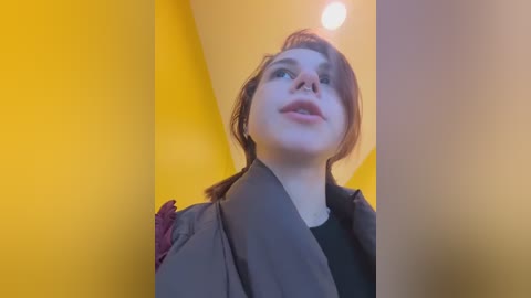 Video of a young woman with light skin, brown hair, and glasses, wearing a gray jacket over a black top, standing against a yellow wall with a bright ceiling light.
