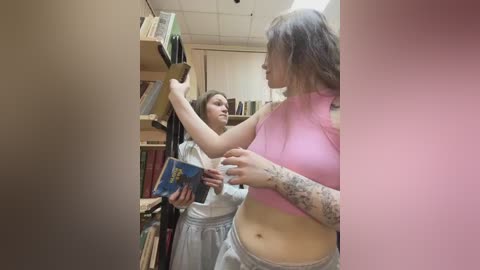 Video of two women in a library: one in a pink crop top, the other in a white dress. They're browsing bookshelves, with the pink-top woman's tattooed arm visible.