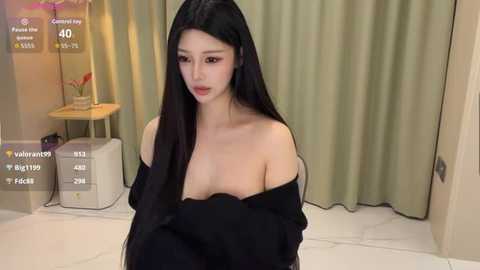 Video of a fair-skinned Asian woman with long black hair, wearing a black off-shoulder dress, sitting on a white floor in a room with beige curtains and a lamp.