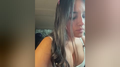Video of a young woman with long, wavy dark hair, wearing a light gray tank top, revealing cleavage. She has a light skin tone and a neutral expression. Background is blurry, indoor setting.