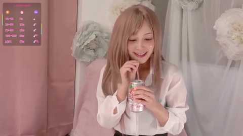 Video of a young Asian girl with straight, light brown hair, wearing a white blouse, sipping from a can while smiling. Pink and white decor background.