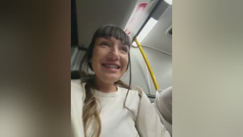 Video of a smiling woman with long brown hair in a white coat, standing on a train. The background features train seats and a yellow pole.