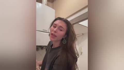 Video of a young Asian woman with long brown hair, wearing a grey shirt and dangling earrings, standing in a beige-tiled kitchen with white appliances.