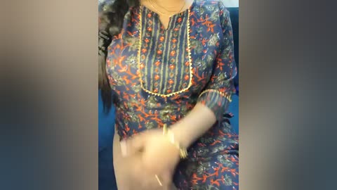 A video of a woman wearing a colorful, embroidered blue kurta with red and gold floral patterns, standing indoors with a blurred background.