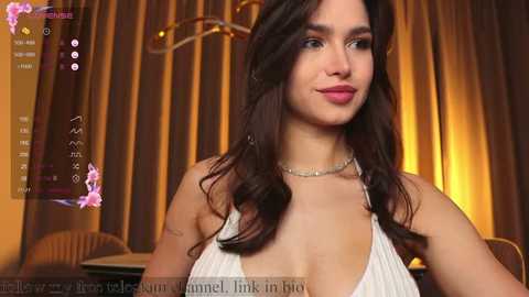 Media: Video of a young woman with wavy dark hair, wearing a white halter top, in a dimly lit room with golden curtains and chandelier.